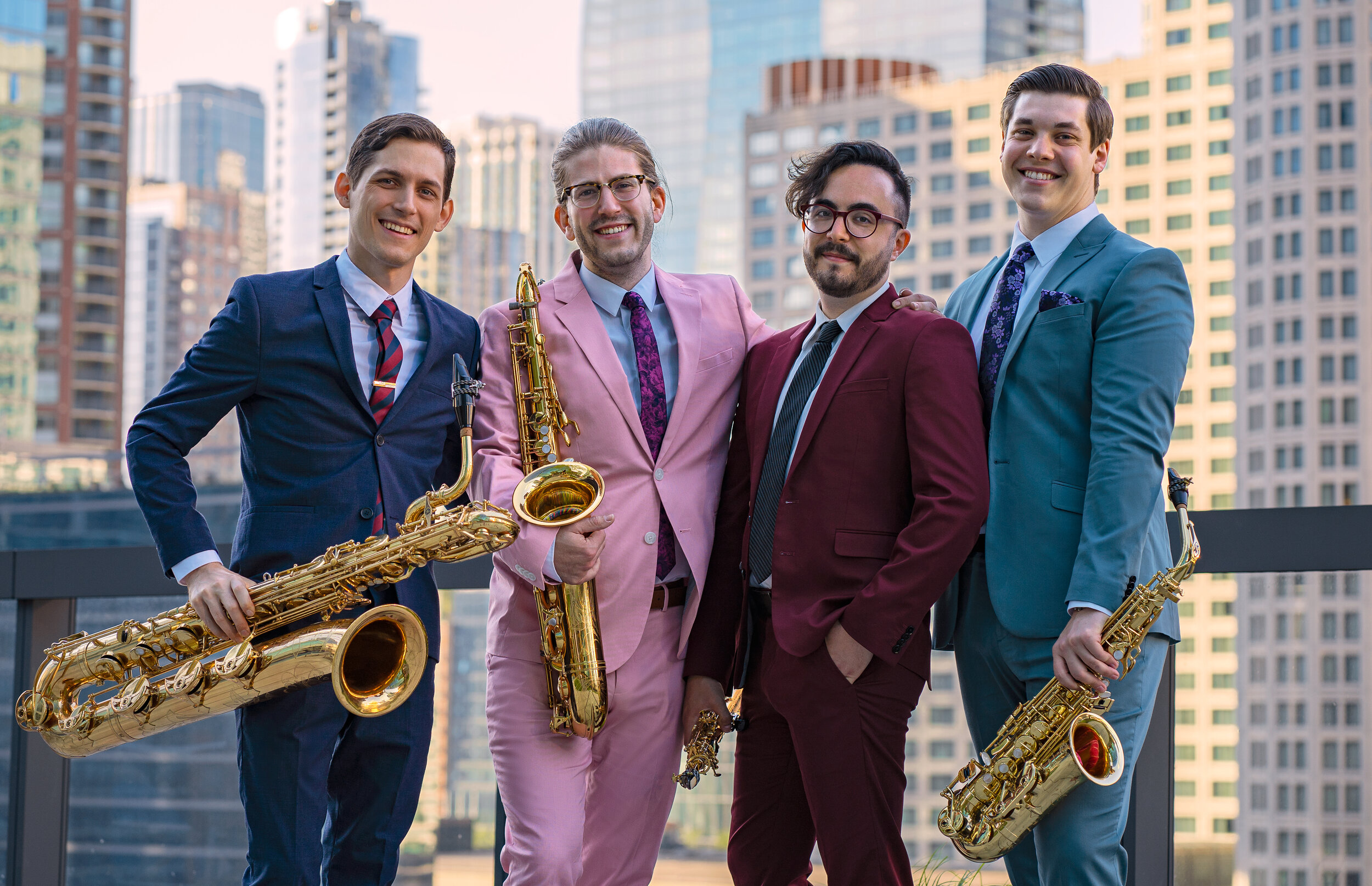 for members of the ~Nois saxophone ensemble. Each member is standing and holding a saxophone, with a blurred city skyline in the background.
