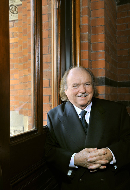 John Perry sitting inside in front of a window, smiling with hands clasped