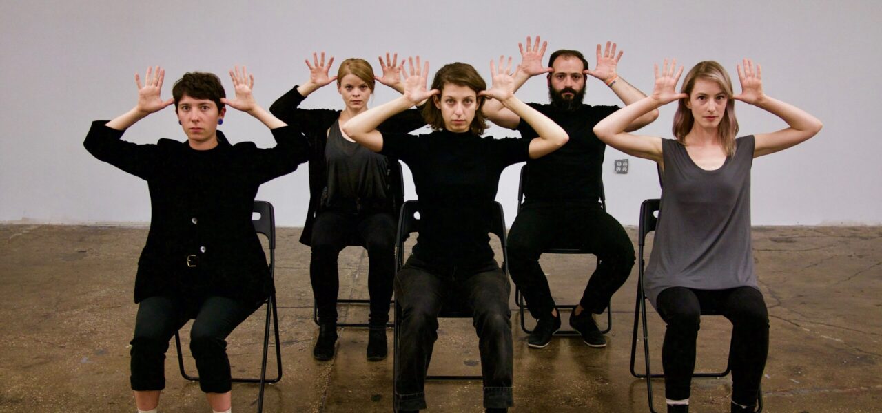 A group of people wearing black ensemble and sitting on chairs with their hands up